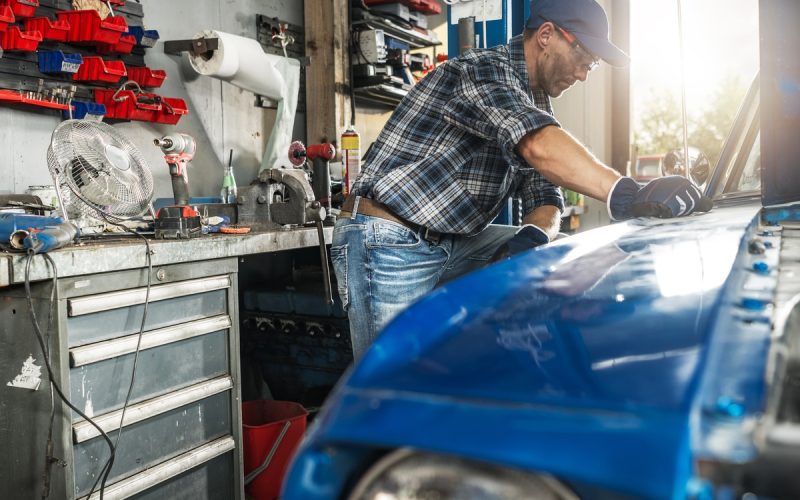 A person wearing a cap and gloves leaning over a blue car with its hood open inside a garage with tools.