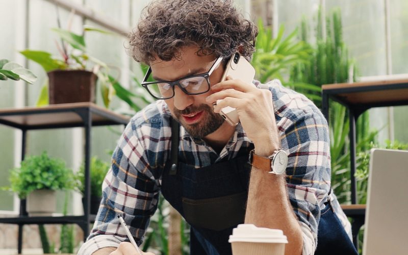 A man wearing glasses and an apron while talking on a phone and writing notes beside a laptop in a plant-filled workspace.
