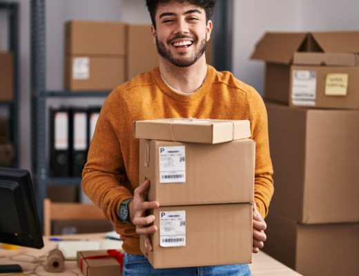 A close-up of a man holding a stack of shipping boxes in a room with shelves, packages, and packing supplies.