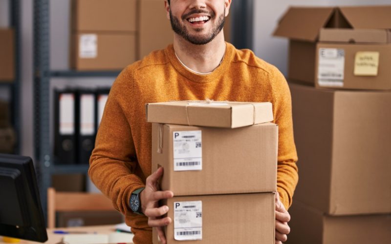 A close-up of a man holding a stack of shipping boxes in a room with shelves, packages, and packing supplies.