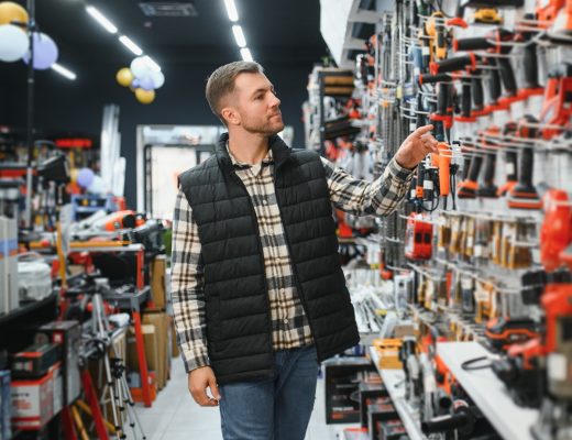 A man in a vest browsing a hardware store aisle while reaching toward a wall display filled with power tools.