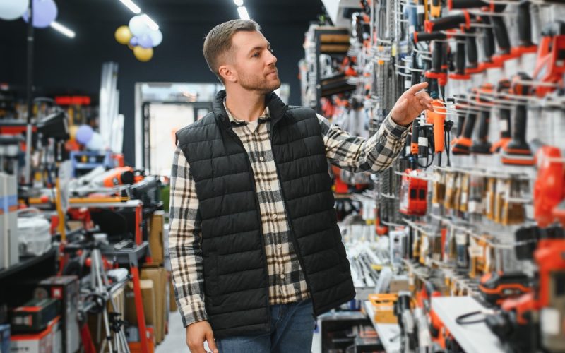 A man in a vest browsing a hardware store aisle while reaching toward a wall display filled with power tools.