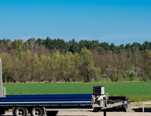 A blue flatbed trailer with two upright loading ramps is parked beside a field. Trees and a blue sky are in the background.