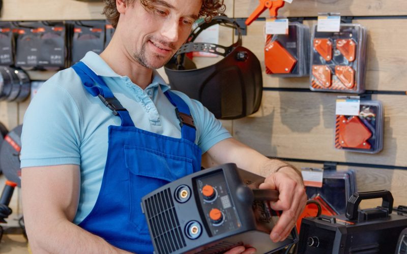 A close-up of a person in work overalls examining a welding machine on a shelf inside a hardware store aisle.