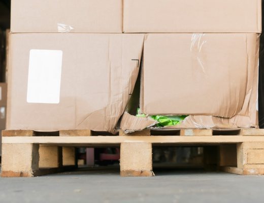 A ground-level close-up of a wooden pallet with stacks of damaged cardboard boxes on it inside a warehouse.