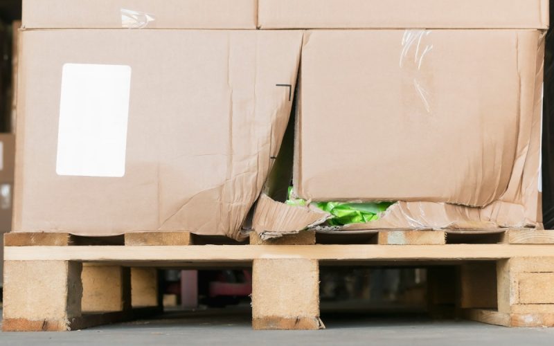 A ground-level close-up of a wooden pallet with stacks of damaged cardboard boxes on it inside a warehouse.
