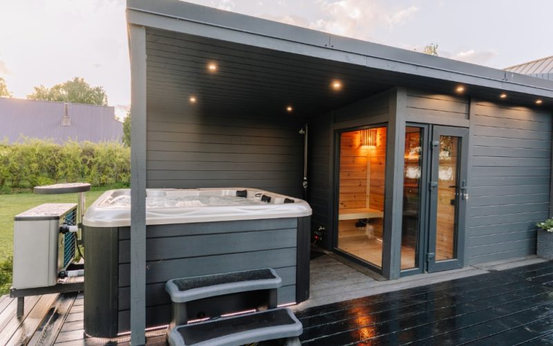 The backyard of a home with a raised deck that includes a hot tub covered by an adjoining cabin and sauna.