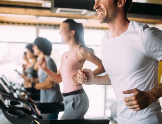 Smiling man runs on a treadmill in a bright gym while several other people exercise on treadmills behind him.