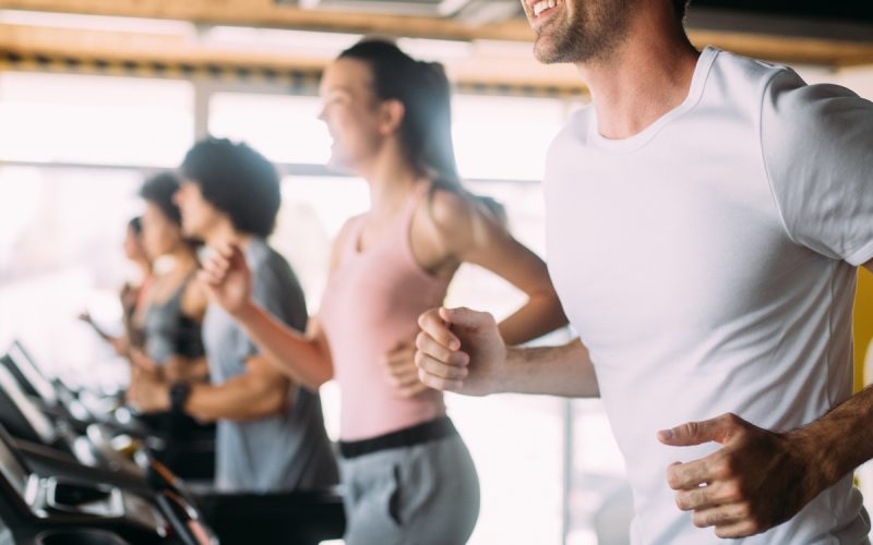 Smiling man runs on a treadmill in a bright gym while several other people exercise on treadmills behind him.